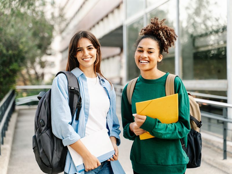 Two students smiling at The Main On University, standing outdoors with backpacks and notebooks, with glass buildings and trees in the background.gmu-dorms-off-campus-housing