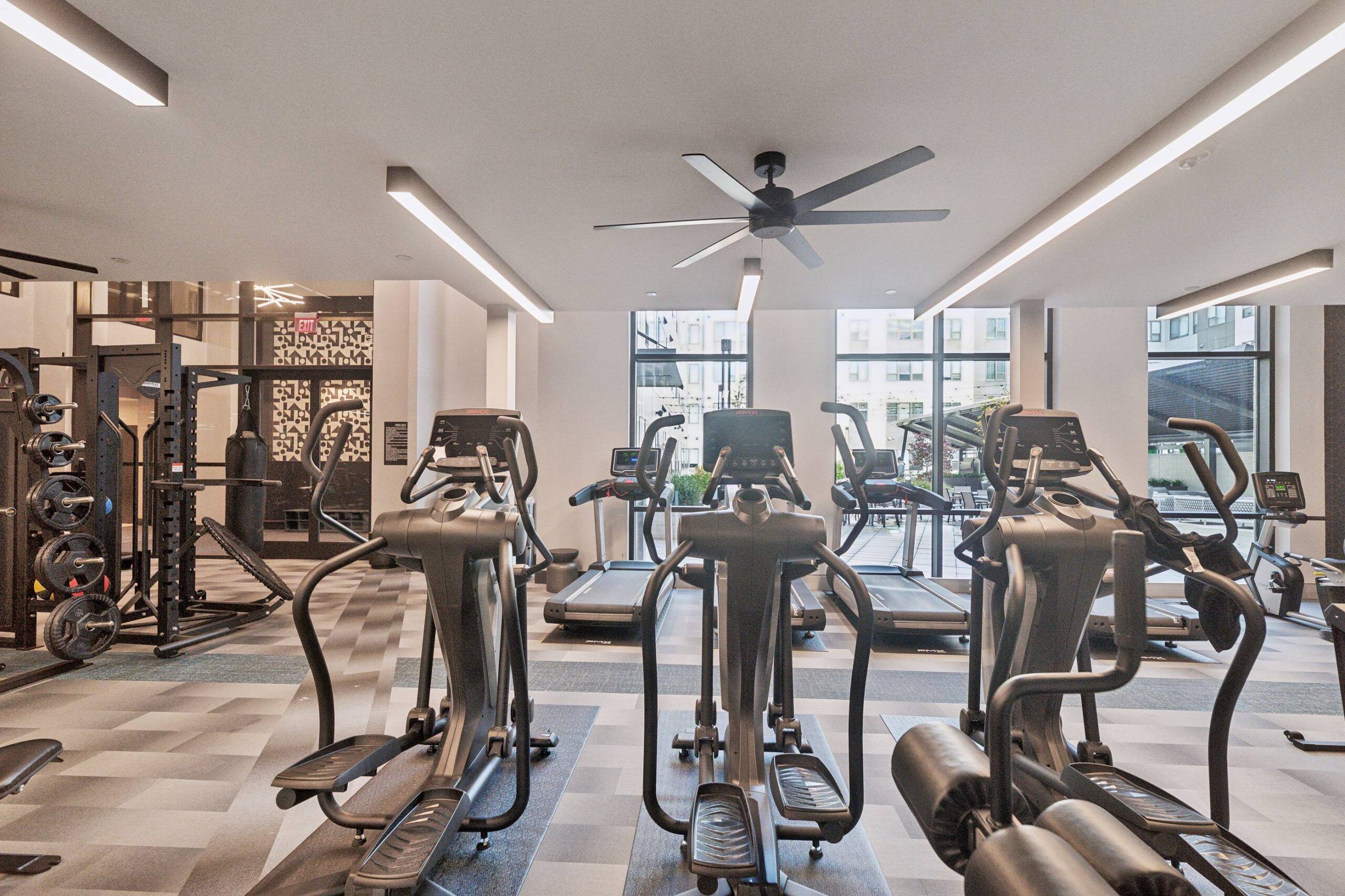 The Main On University features a modern gym with treadmills, ellipticals, weights, large windows, and ceiling fans on a gray checkered floor.
